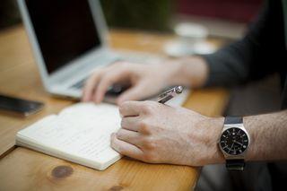 man's hand writing on a pad next to a laptop
