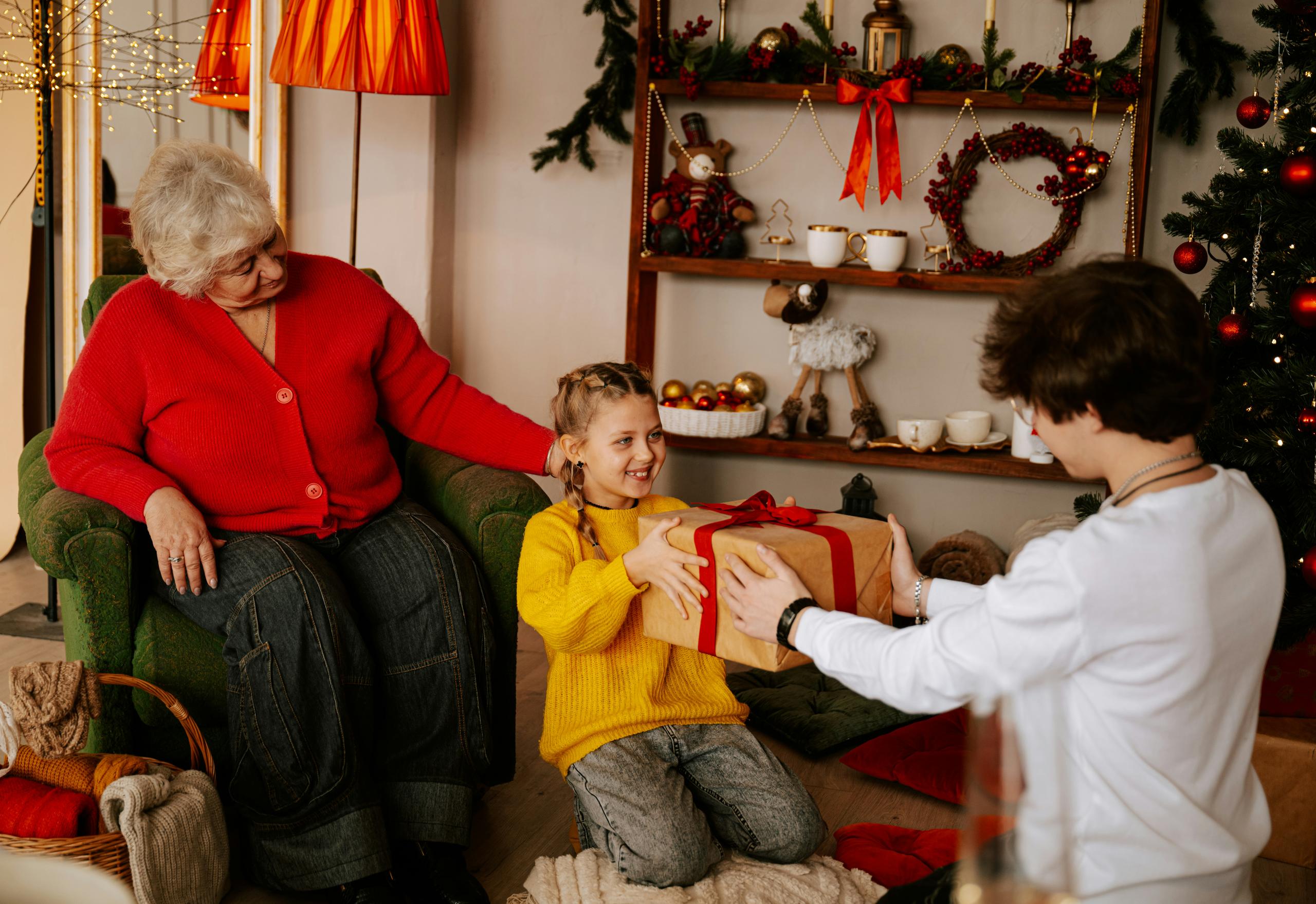 An elderly woman in a red sweater sits in an armchair smiling as a young girl in a yellow sweater kneels on the floor receiving a wrapped gift with a red ribbon from an older child, in a cozy living room decorated with Christmas ornaments, garlands, wreaths, and warm lights.